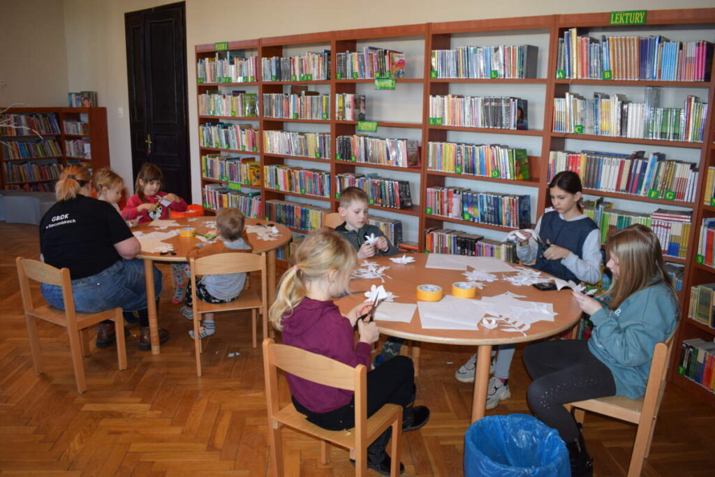 Przy stołach siedzą dzieci i wycinają z papieru elementy dekoracji. W tle stoją półki z książkami.