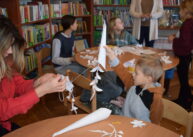 Dzieci siedzą przy stołach. Jedno dziecko podnosi do góry swoją pracę – zrobiony z papieru sopel lodu.