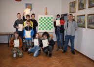 Grupa szachistów pozuje do zdjęcia. Ustawieni są wokół szachownicy zawieszonej na sztaludze. W rękach trzymają dyplomy.