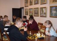 Przy stole zawodnicy grają w szachy. Z tyłu na ścianie wiszą obrazy.