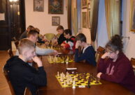 Przy stołach ustawionych przy oknie zawodnicy grają w szachy.
