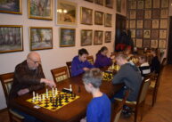 Przy stole zawodnicy grają w szachy. Z tyłu na ścianie wiszą obrazy.