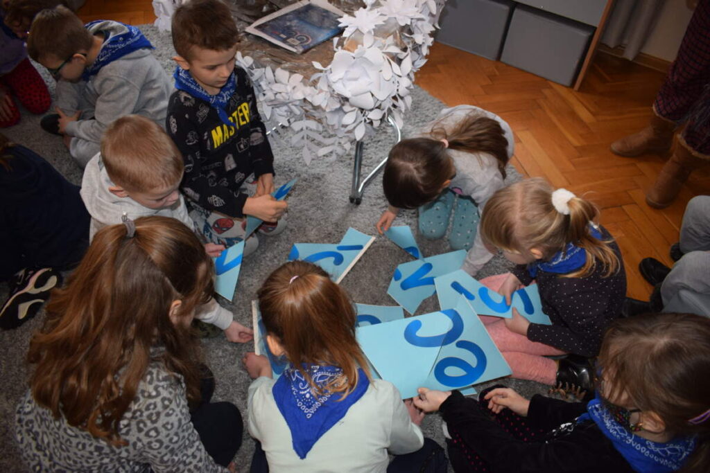 Siedząca na dywanie grupka dzieci układa z zadrukowanych kartek fragment tekstu.