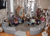 Wokół okrągłego dywanu ustawione są pufy. Dzieci siedzą na dywanie w dwóch grupach. Nad głowami wiszą wykonane z papieru ozdoby.