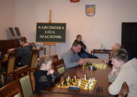 Przy stole para zawodników gra w szachy. W tle widać mężczyznę siedzącego przy stole.