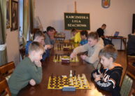 Przy rzędzie stołów ustawionych przy oknie zawodnicy grają w szachy. W tle widoczna tablica z napisem KARCZMISKA LIGA SZACHOWA.