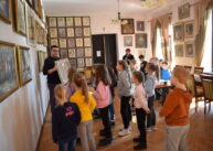 Grupa dzieci stoi wokół dorosłego mężczyzny, który pokazuje dzieciom obraz. Na ścianach wiszą oprawione rysunki i obrazy.