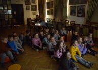 Dzieci siedzą na podłodze i oglądają film. W pomieszczeniu jest ciemno.