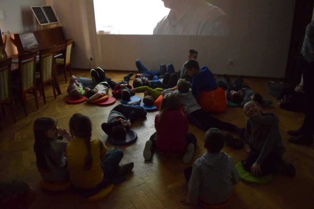 Dzieci siedzą na podłodze i oglądają wyświetlany na ścianie film. W pomieszczeniu jest ciemno.