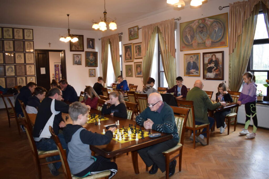 Zdjęcie całej sali. Widać dwa rzędy stołów, przy których trwają rozgrywki szachowe.