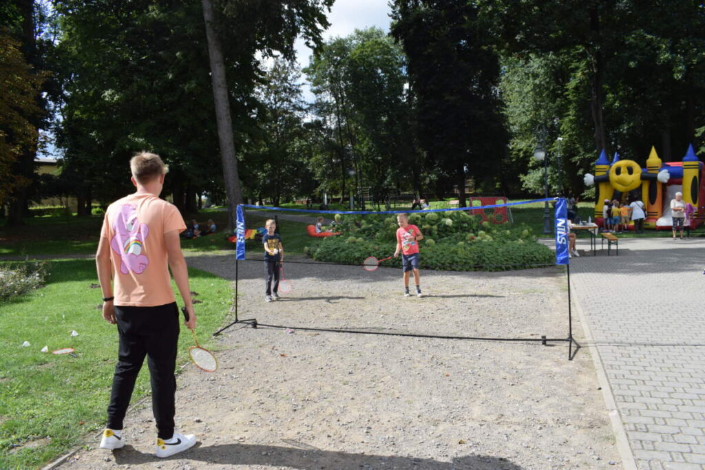 Kilkoro dzieci gra w badmintona. W tle widać dzieci siedzące na pufach.