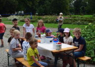 Dzieci siedzą na ławce przy stole przykrytym białym materiałem. Na stole stoją owoce.