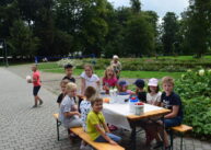 Dzieci siedzą na ławce przy stole przykrytym białym materiałem. Na stole stoją owoce.