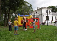Dzieci grają w piłkę. Za dziećmi widoczna jest bramka z naciągniętym czerwonym materiałem, w którym są duże otwory. Z tyłu za bramką widoczny jest dmuchany zamek.