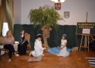 Przy stole siedzi dwoje dzieci i czyta. Kolejna dwójka klęczy na środku sali. W tle dekoracja: sztuczne drzewo z zawieszoną na nim kapliczką; za drzewem ściana z zielonego materiału.