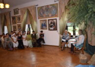 Zdjęcie sali. Dwie kobiety siedzą przy stole i czytają. Pozostali uczestnicy wydarzenia siedzą na krzesłach. Na ścianie wiszą obrazy.