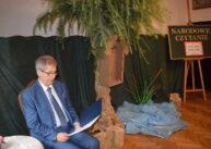 Mężczyzna ubrany w garnitur siedzi na krześle i czyta. Obok niego stoi drzewo z zawieszoną kapliczką. Obok drzewa leży niebieska folia a za nią stoi sztaluga z tablicą z napisem NARODOWE CZYTANIE.