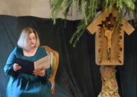Kobieta ubrana w zieloną sukienkę siedzi na krześle i czyta. Obok niej stoi drzewo z zawieszoną kapliczką.