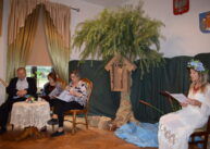 Na tle dekoracji z zielonego materiału przy stole siedzą ubrani w różne stroje ludzie i czytają. Na środku stoi drzewo z zawieszoną na nim kapliczką.