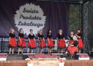 Na scenie stoją ubrane w czarne bluzki i czerwone spódnice kobiety. W tle sceny jest fioletowy materiał z napisem ŚWIĘTO PRODUKTU LOKALNEGO.