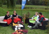 Na ustawionych na trawie kanapach siedzą czworo ludzi i rozmawiają ze sobą. Przed nimi na stole stoją różne potrawy.