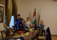 Na końcu rzędu stołów stoi grupka dzieci. Na stole ustawione są plansze z szachami. Za dziećmi widoczne są trzy flagi.
