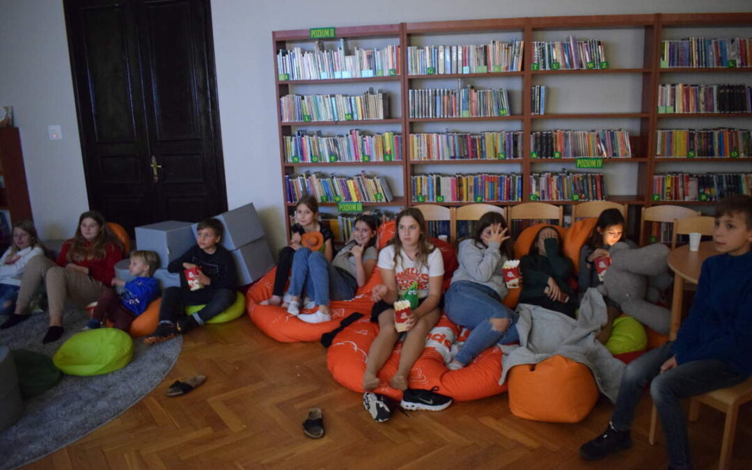 Dzieci siedzą na pufach i oglądają film. Za dziećmi pułki z książkami.