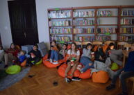 Dzieci siedzą na pufach i oglądają film. Za dziećmi pułki z książkami.