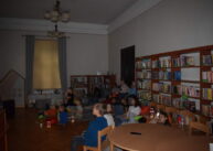 Dzieci siedzą i oglądają film. Na pierwszym planie widoczny fragment okrągłego stołu. W tle widoczne są półki z książkami.
