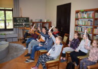 Dzieci siedzą na, ustawionych w dwóch rzędach, krzesłach. Niektórzy mają podniesione ręce i trzymają kartki z literami. W tle widać półki z książkami.