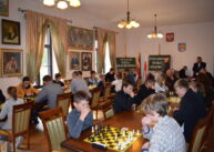 Przy ustawionych równolegle rzędach stołów siedzą szachiści i grają w szachy.