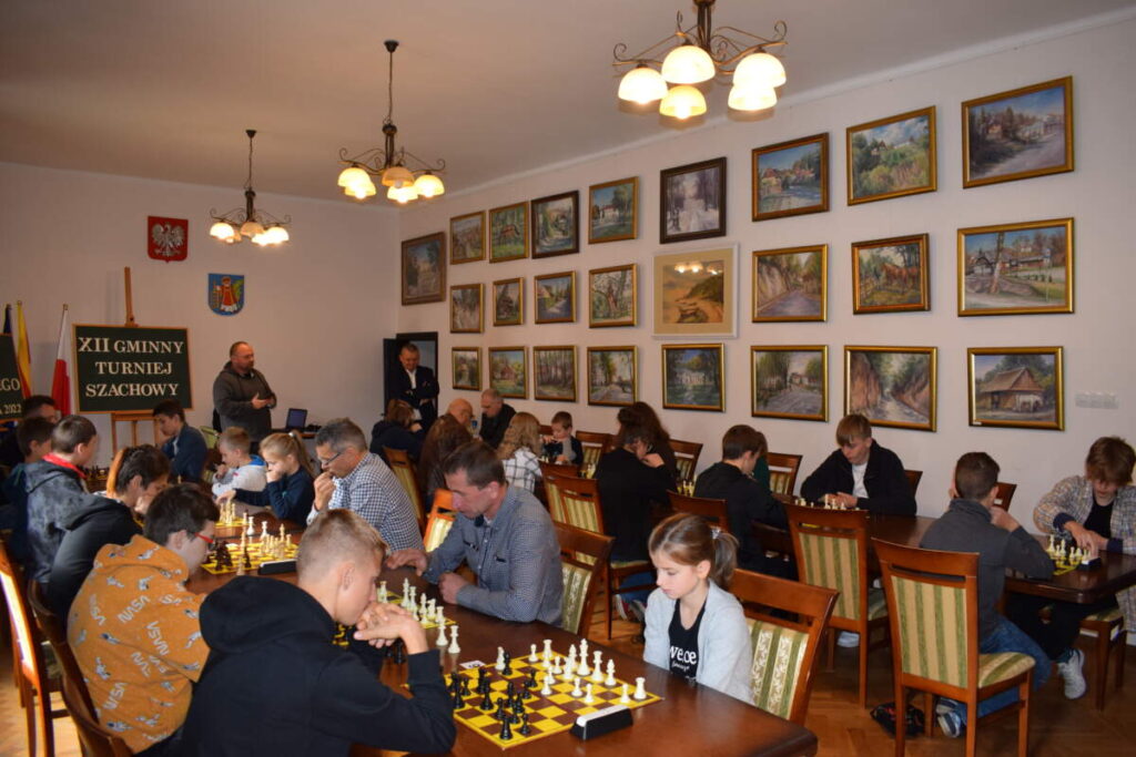 Przy ustawionych równolegle rzędach stołów siedzą szachiści i grają w szachy. w tle na ścianach wiszą obrazy.