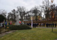 Grupy ludzi stoją wokół pomnika. Pośrodku widoczny jest biały maszt.