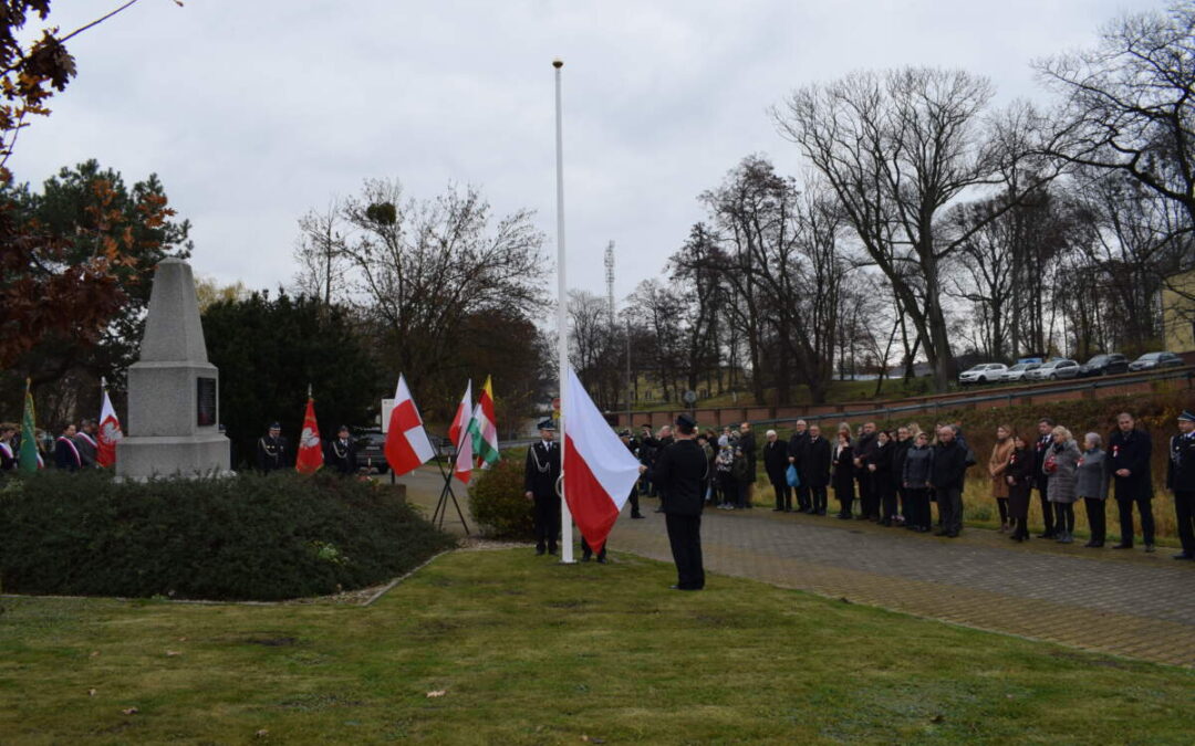 Grupy ludzi stoją wokół pomnika. Pośrodku widoczny jest biały maszt, do którego zamocowana jest biało czerwona flaga. Stojący obok mężczyzna unosi róg flagi do góry.