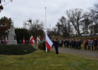Grupy ludzi stoją wokół pomnika. Pośrodku widoczny jest biały maszt, do którego zamocowana jest biało czerwona flaga. Stojący obok mężczyzna unosi róg flagi do góry.