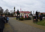Grupy ludzi stoją wokół pomnika. Pośrodku widoczny jest biały maszt z biało czerwoną flagą.