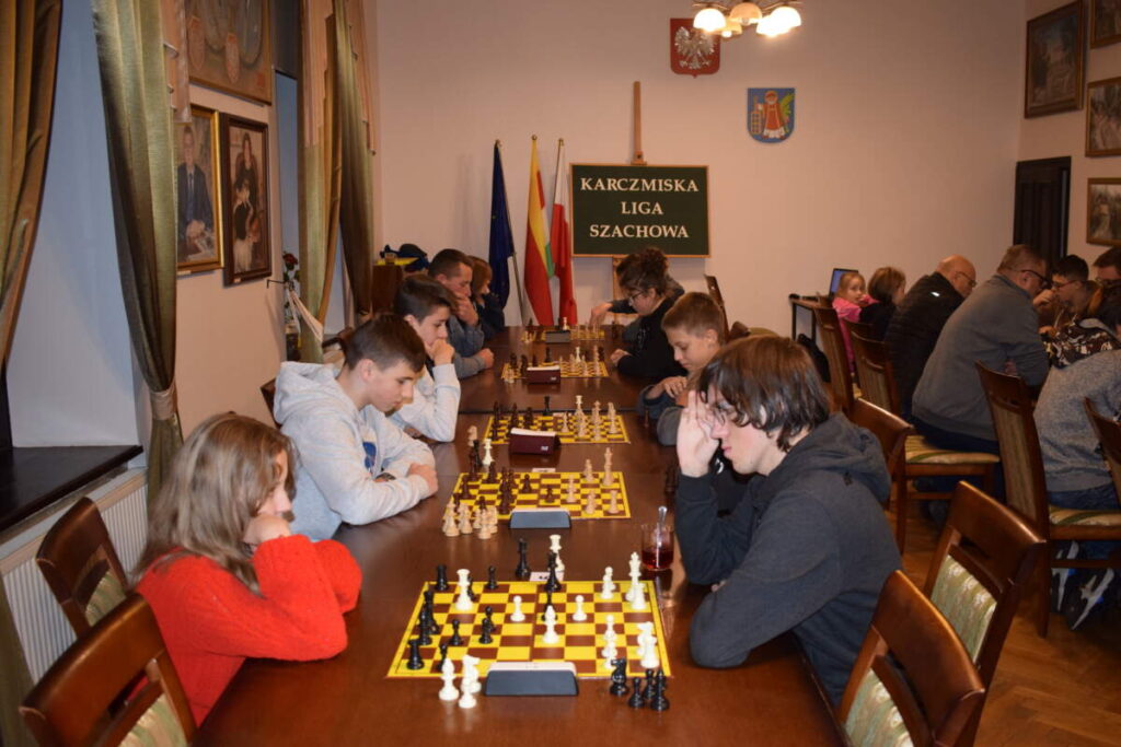 Przy ustawionych w rzędzie stołach siedzą zawodnicy i grają w szachy. W tle tablica z napisem KARCZMISKA LIGA SZACHOWA.