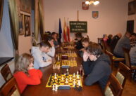 Przy ustawionych w rzędzie stołach siedzą zawodnicy i grają w szachy. W tle tablica z napisem KARCZMISKA LIGA SZACHOWA.