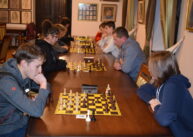 Przy ustawionych w rzędzie stołach siedzą zawodnicy i grają w szachy..