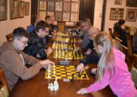 Przy ustawionych w rzędzie stołach siedzą zawodnicy i grają w szachy..