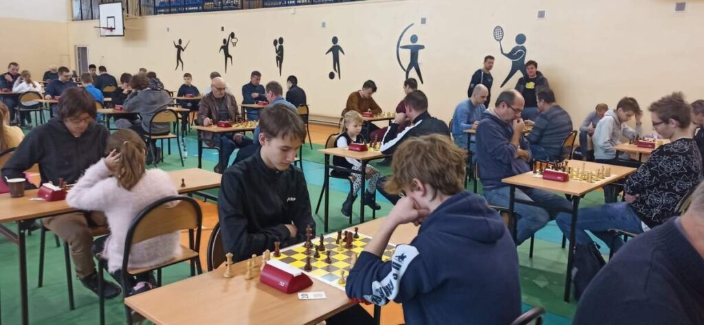Na pierwszym planie dwie osoby siedzi przy stole na przeciw siebie i gra w szachy. W tle pozostali zawodnicy grają w szachy.