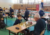 Na pierwszym planie dwie osoby siedzi przy stole na przeciw siebie i gra w szachy. W tle pozostali zawodnicy grają w szachy.