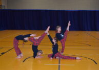 Dzieci wykonują figury akrobatyczne. Ubrane w czarne bluzki i czerwone spodnie w kratkę.