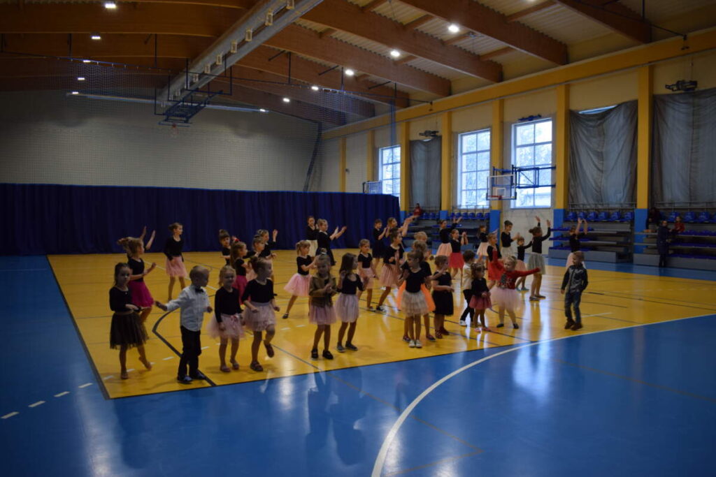 Widok na cała halę sportową. Grupa, kolorowo ubranych dzieci tańczy.