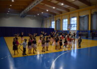 Widok na cała halę sportową. Grupa, kolorowo ubranych dzieci tańczy.