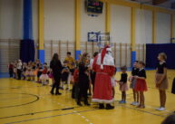 Na zdjęciu widoczna grupa dzieci. Stoją w rzędzie. Ubrany w biało czerwony strój m0ikołaj rozdaje dzieciom prezenty.