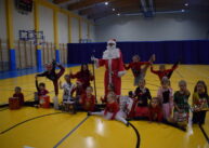 Na zdjęciu widoczna pozująca do zdjęcia grupka dzieci, za nimi ubrany w biało czerwony strój mikołaj.