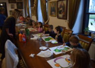 Przy stole siedzą dzieci i rysują. Na stole leżą kartki i materiały plastyczne.