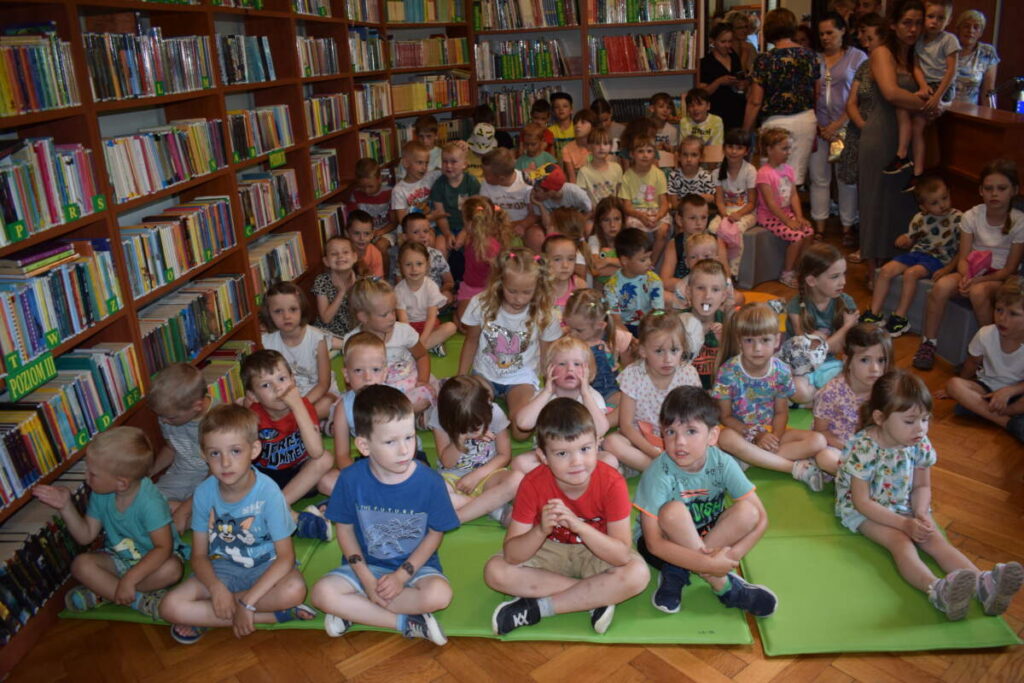 Na rozłożonych na podłodze materacach siedzą dzieci. W tle na półkach stoją książki.