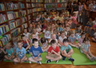 Na rozłożonych na podłodze materacach siedzą dzieci. W tle na półkach stoją książki.